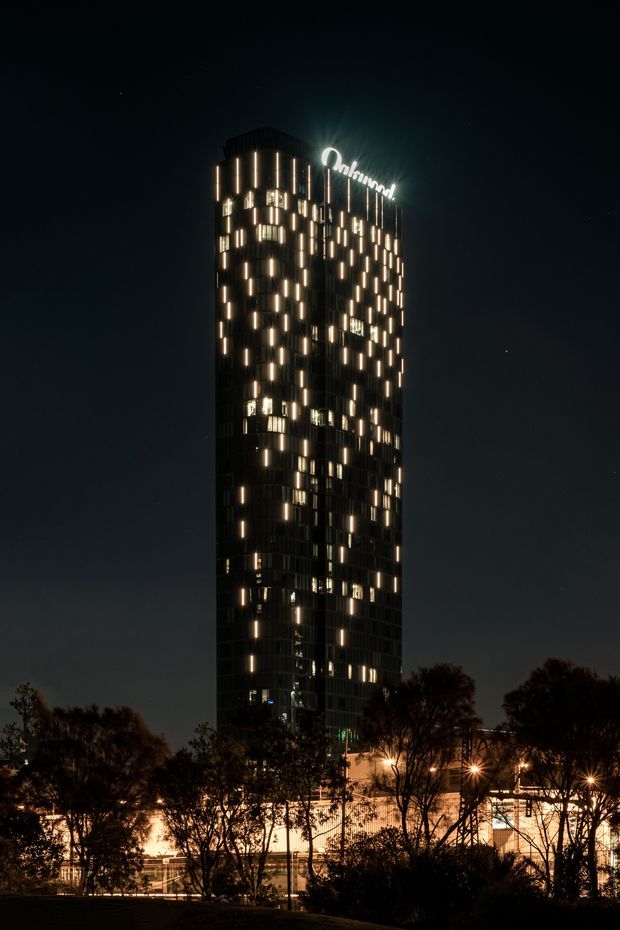 Night-time exterior photograph of a tall, narrow commercial high-rise tower with a dark glazed facade punctuated by numerous warm illuminated vertical windows. A bright rooftop sign is visible, with trees and low-rise illuminated structures in the foreground. The image has a dramatic urban architectural photography style.