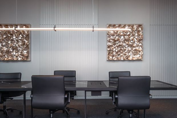 A contemporary office meeting room with a long dark conference table surrounded by black leather office chairs. The room features a vertically ribbed grey wall finish, dark carpet flooring, slim linear pendant lighting, and two framed textured metallic artworks.