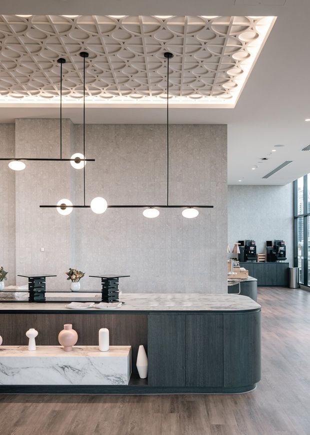 A refined contemporary hospitality or commercial reception and lounge space with a large dark timber-clad island counter, pale marble worktop and display ledge, textured grey wall panels, and sculptural black-and-white pendant lighting. A decorative recessed ceiling features a geometric circular-patterned panel with concealed illumination. Coffee equipment and tall windows are visible in the background, while the space uses warm timber flooring and restrained neutral tones.