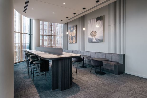 A contemporary commercial dining or breakout area with a long communal table, black bar stools, upholstered banquette seating, and small round café tables. Floor-to-ceiling glazing overlooks an urban high-rise setting, while vertical wall panelling, pendant lights, recessed ceiling lighting, and muted blue-grey finishes create a refined modern interior.
