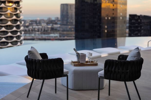 A refined high-rise apartment lounge with two black upholstered armchairs facing a low white ottoman coffee table. Floor-to-ceiling glazing overlooks a dense urban skyline and a rooftop or terrace beyond. The room has pale stone or tile flooring, a restrained neutral palette, and softly rounded contemporary furniture.