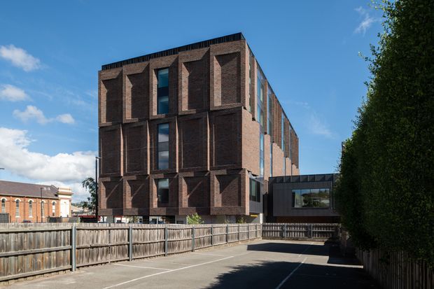 Exterior view of a contemporary multi-storey brick building with a strongly articulated facade of deep vertical window recesses and projecting brick piers. A glazed connector links the main building to a darker low-rise volume, while a paved driveway and timber fencing occupy the foreground. The image also shows an adjacent traditional brick building, mature screening vegetation, and a clear blue sky.
