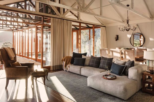 A bright, spacious open-plan living room with exposed white-painted roof trusses and beams, extensive timber-framed glazing, pale vertical wall panelling, and polished dark timber flooring. A large neutral modular sectional sofa anchors the room, with leather lounge chairs, a dining area, linen curtains, a round mirror, and a decorative chandelier. Sunlight fills the room through the glazed corner, creating a warm, relaxed interior.