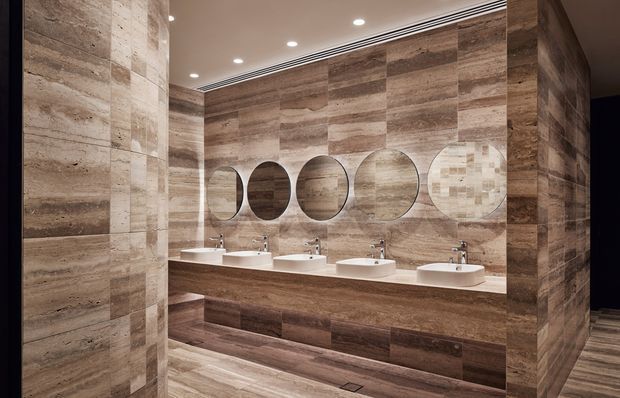 A refined contemporary bathroom vanity area clad extensively in brown, wood-look stone or tile. A long floating double-or-more vanity with multiple white vessel basins and chrome tapware sits beneath a large mirror composed of overlapping circular forms. Recessed ceiling downlights and linear air-conditioning grilles provide clean modern detailing, while the continuous wall, floor, and joinery finishes create a warm, cohesive interior.
