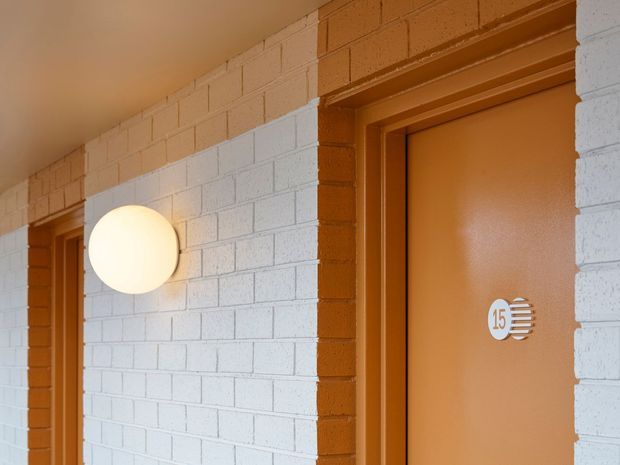 Close-up view of a corridor or building entrance with white-painted concrete block walls, warm orange-brown painted door frames and doors, and a round wall-mounted globe light. The visible door is marked with the number 15, and a second doorway is partially visible at left. The image emphasizes the facade and entry doors rather than an interior room.
