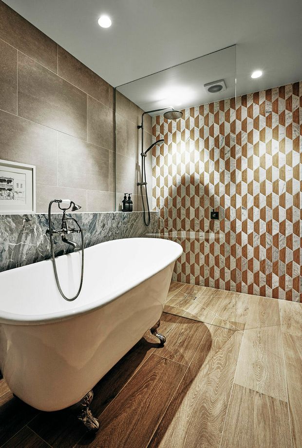 A refined bathroom featuring a freestanding white clawfoot bathtub, wall-mounted bath and shower fixtures, a large frameless mirror, recessed ceiling lights, marble-look lower wall tiles, large taupe wall tiles, and a striking geometric patterned tile shower wall in warm brown, cream, and grey tones. The floor combines warm wood-look and pale stone-look tiles.