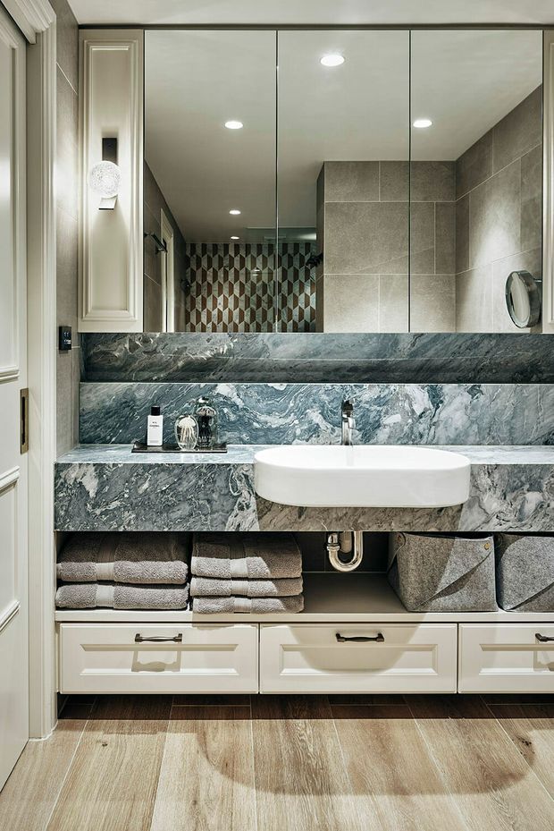 A refined bathroom vanity area with a white vessel basin set into a dramatic grey-green veined stone benchtop and backsplash. Mirrored wall panels expand the space, while cream framed cabinetry, integrated open shelving with folded towels and fabric baskets, recessed ceiling lights, and large-format grey wall tiles create a polished, traditional-contemporary look. Timber-look flooring adds warmth.