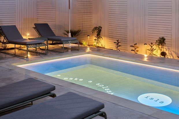 A modern outdoor pool area photographed at dusk, featuring a rectangular illuminated swimming pool with in-water projected text, pale paving, dark sun loungers, warm landscape lighting, and a slatted vertical privacy wall with integrated planting.