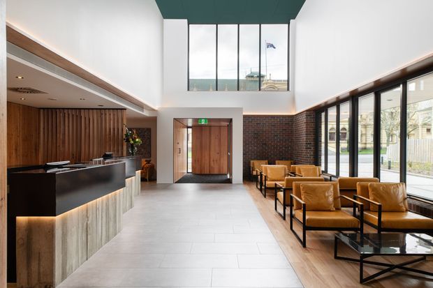 A refined contemporary reception lobby with a double-height ceiling and large upper-level glazing. The space features a dark reception desk with integrated warm lighting, vertical timber wall panelling, a pale tiled floor, exposed red brick, extensive black-framed windows, and a row of tan leather lounge chairs. A timber entrance door sits beneath a green exit sign, with an urban streetscape visible through the glazing.