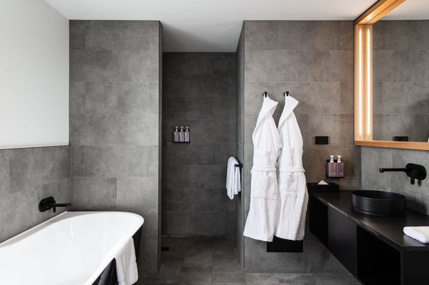 Contemporary bathroom featuring a freestanding white bathtub, dark round vessel basin, wall-mounted black tapware, charcoal grey tiled walls and floor, and black floating vanity cabinetry. White robes hang beside a walk-in shower area, while a large timber-framed mirror with integrated vertical lighting adds warmth.