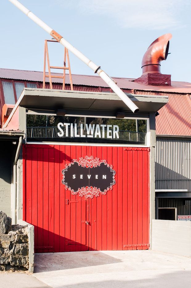 Exterior view of an industrial-style building entrance with vivid red timber garage or loading doors, corrugated metal roofing and cladding, exposed ductwork, and prominent signage reading “STILLWATER” and “SEVEN.” A small stone wall and concrete driveway are visible in the foreground.