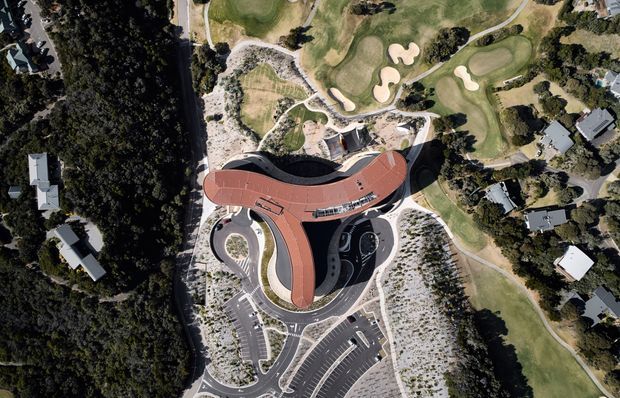 Aerial top-down view of a large contemporary building with a distinctive curved, branching red roof, surrounded by extensive paved circulation areas, parking, landscaped grounds, dense vegetation, and an adjacent golf course. The image primarily documents the building's overall site plan and roof form.
