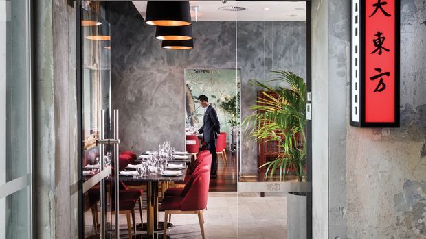 View through glass doors into a refined contemporary restaurant dining area. The space features a long set dining table with white place settings, upholstered red dining chairs, black pendant lights, a textured grey feature wall, indoor palms, and a red vertical sign with Asian characters. Exposed textured surfaces and dark metal-framed glazing add an urban, polished character.
