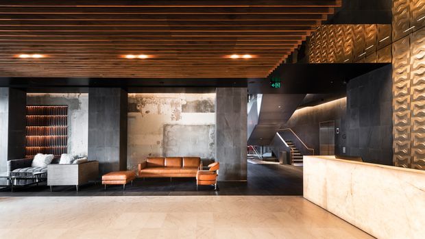 A dramatic, refined commercial lobby or reception area with a dark industrial-inspired fit-out. The space features a large pale stone reception desk, a leather sofa and lounge seating, blackened and textured wall finishes, a timber slatted ceiling with integrated lighting, and a stairway leading deeper into the building.