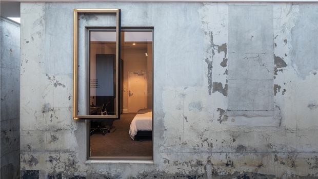 Exterior view of a weathered concrete or plaster facade with a narrow window opening. The projecting timber-framed window is open, revealing a warm-toned bedroom interior with a visible bed, desk or chair, and built-in cabinetry. The facade has distressed, patchy grey surfaces and a strong contrast between the rugged exterior and refined interior.