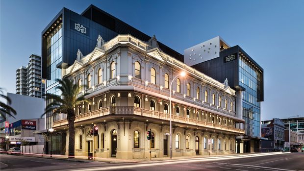Street-level dusk view of a historic ornate multi-storey commercial building with cream masonry, classical detailing, arched windows, balconies and a corner entrance, integrated with a contrasting black contemporary tower and glazed volumes behind it. The building occupies a prominent urban intersection with surrounding high-rise development and illuminated architectural lighting.