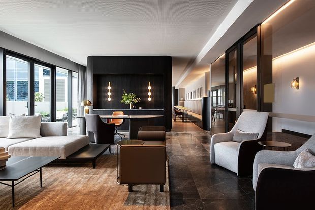 A sophisticated open-plan contemporary interior combining a lounge, dining area and linear kitchen or service zone. The room features floor-to-ceiling black-framed glazing, a dark ribbed feature wall with integrated lighting, dark stone-look flooring, warm timber and bronze-toned joinery, layered recessed and wall lighting, and a restrained palette of cream, grey, black and brown. A large textured rug anchors the seating area, while sculptural upholstered chairs and sofas create a refined hospitality-like atmosphere.