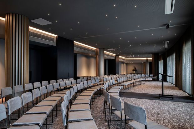 A large contemporary auditorium or event hall arranged with multiple curved rows of light upholstered chairs facing a presentation area. The room features dark acoustic ceilings with recessed spotlights, integrated linear lighting, black wall panels, vertical timber slat columns, floor-to-ceiling curtains, and patterned carpet. A lectern stands at the front-right of the space.
