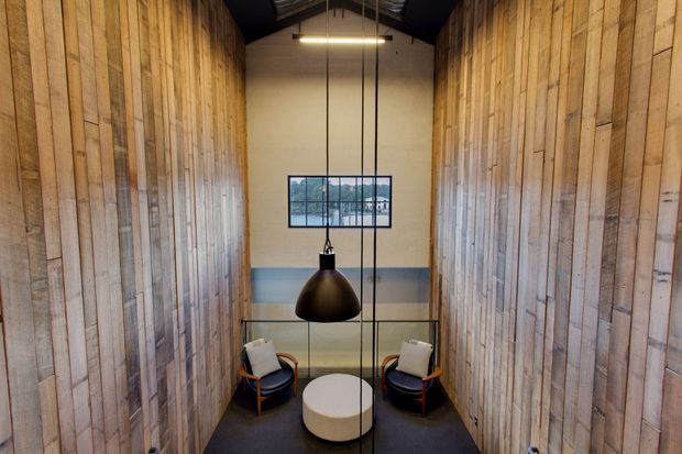 A narrow, tall interior lounge or waiting area with warm reclaimed timber wall panelling on both sides, a dark exposed pitched roof structure, and a central black pendant light. Two upholstered lounge chairs and a round white coffee table sit at the far end, with a black-framed window and glazed balustrade adding depth. The palette is warm timber, charcoal, cream and muted blue-grey, creating a refined industrial atmosphere.