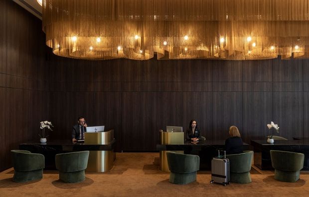 A sophisticated hotel or commercial reception area with two reception desks set against dark vertical wood panelling. Deep green upholstered lounge chairs sit in front of the desks, while a large suspended chandelier made from dense vertical amber-toned strands provides warm illumination. The space has a dark, luxurious palette and timber flooring.