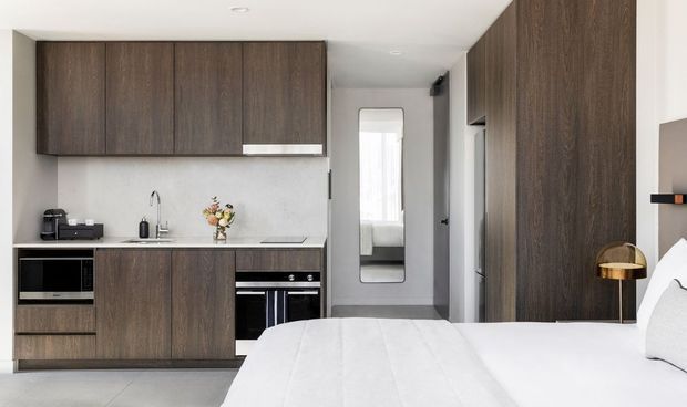 Compact open-plan studio interior combining a kitchenette and sleeping area. Dark wood cabinetry runs along the kitchen wall, paired with a light stone benchtop, integrated appliances, pale grey walls, and a white bed in the foreground. A full-height dark wood wardrobe or storage wall occupies the right side, with a narrow hallway and mirror leading deeper into the apartment. The look is clean, contemporary, and space-efficient.