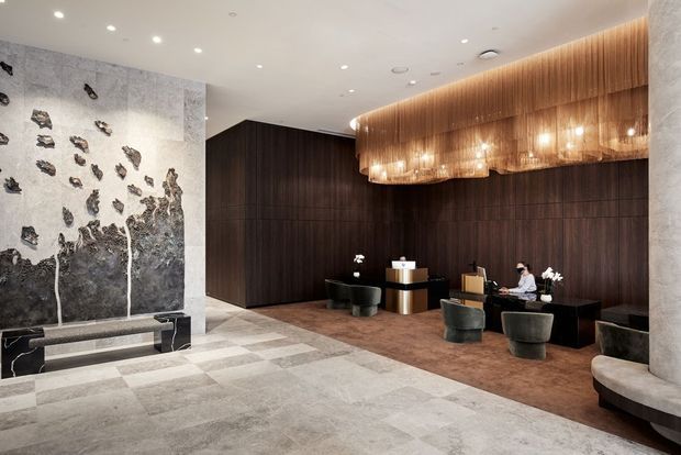 A sophisticated hotel or commercial lobby with dark timber wall panelling, a long reception desk, sculptural grey seating, and a large decorative stone feature wall. The space is illuminated by a dramatic suspended timber-toned chandelier with integrated pendant lights, while large-format pale stone flooring creates a refined, luxurious atmosphere.