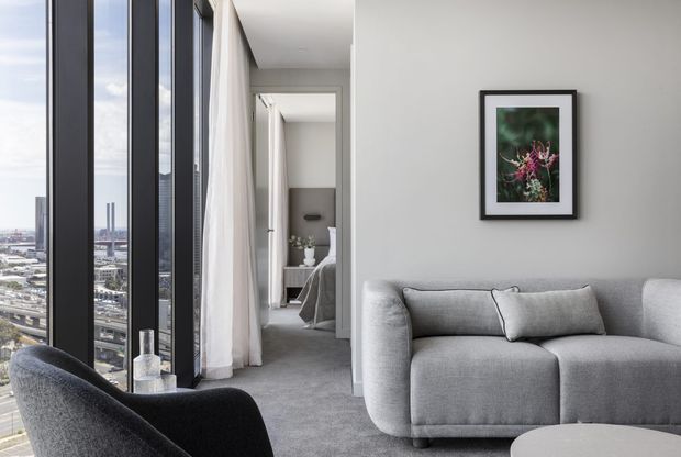 A bright contemporary apartment living room with a large light-grey rounded sofa, black upholstered lounge chair, pale grey carpet, white walls, and a framed botanical artwork. Floor-to-ceiling dark-framed glazing provides expansive city views, while a hallway and partially visible bedroom appear beyond the living area.