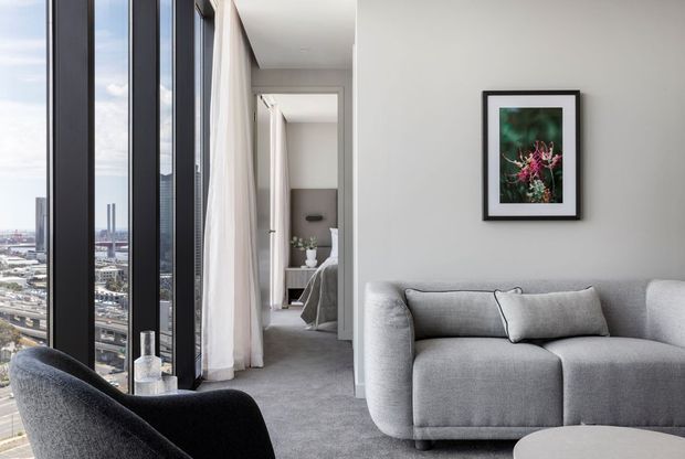 A bright, refined high-rise living room with floor-to-ceiling black-framed glazing overlooking an urban cityscape. The room features a large light-grey curved sofa, dark upholstered lounge chair, grey carpet, white walls, sheer curtains, and a black-framed botanical artwork. A bedroom or sleeping area is visible through the open doorway in the background.