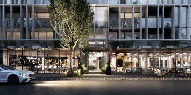 Street-level architectural rendering of a contemporary multi-storey mixed-use building. The facade combines a regular concrete structural grid with extensive floor-to-ceiling glazing, recessed balconies, and planted terraces. The transparent ground floor contains brightly lit commercial interiors, including what appears to be a cafe or restaurant with outdoor seating. A landscaped street tree and entry planting sit in front of the building.