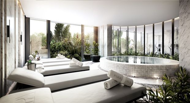 Luxurious contemporary spa-like bathroom with a large circular freestanding soaking tub, white upholstered loungers, floor-to-ceiling curved glazing, marble-clad walls, and abundant indoor planting. The bright, refined space opens visually to lush greenery outside.