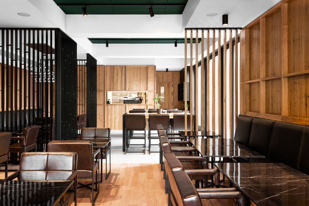 Contemporary hospitality interior with a dining and lounge area arranged around a compact bar or service counter. The space features extensive warm timber wall panelling and built-in cabinetry, black metal screens and framing, dark marble-topped tables, leather upholstered seating, and a bright open ceiling with dark green inset ceiling panels and track lighting.