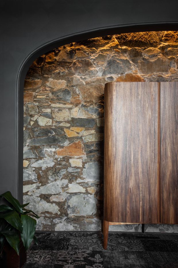A moody interior detail featuring a tall, rounded-top black-painted arch framing an exposed rough stone wall. A large mid-to-dark timber cabinet or sideboard with vertical grain stands against the stone, with slender timber legs and dark hardware. Patterned dark carpet covers the floor, and a leafy indoor plant sits at the lower left. Warm concealed lighting highlights the stonework and contrasts with the predominantly dark palette.