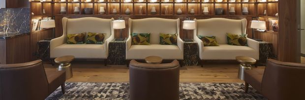 A sophisticated lounge or reception area with three cream upholstered sofas arranged against a dramatic backlit timber display wall filled with decorative objects. Dark marble side tables, table lamps, brown leather lounge chairs, and a patterned area rug create a warm, luxurious hotel or commercial interior.
