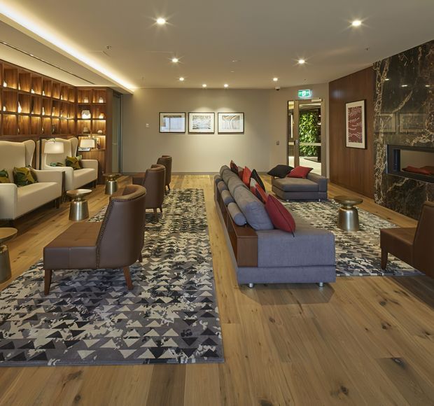 A refined contemporary living room with warm timber flooring, a large sectional sofa, leather lounge chairs, patterned area rugs, illuminated built-in shelving, framed artwork, recessed lighting, and a dramatic dark marble fireplace surround. The room is softly lit and arranged as a comfortable sitting and entertaining space.