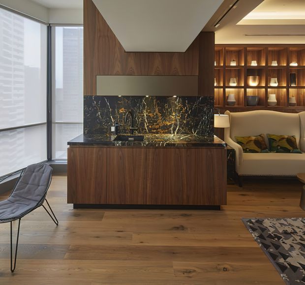 A sophisticated open-plan living area featuring a compact kitchen or bar joinery zone with dark timber cabinetry, a dramatic dark veined stone splashback and benchtop, and integrated lighting. Warm timber flooring continues into a lounge area with a cream sofa, sculptural display shelving, and expansive corner glazing overlooking urban towers.