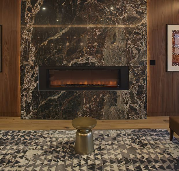 A sophisticated living room feature wall with a linear electric fireplace inset into a dramatic dark veined stone surround. Warm timber wall panelling frames the fireplace, while a patterned geometric rug and small metallic side table add layered contemporary detail.