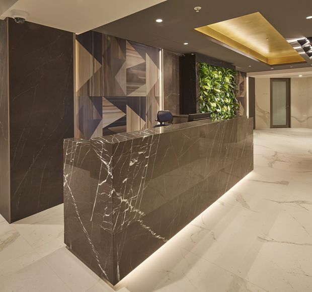 A sophisticated commercial reception lobby featuring a large dark veined stone reception desk with integrated underlighting, matching dark wall panels, a geometric timber-toned feature wall, and a lush vertical garden. Pale veined stone flooring, recessed ceiling lights, and a backlit gold-toned ceiling feature create a luxurious, contemporary atmosphere.