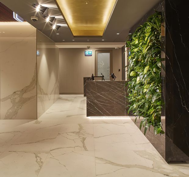 Luxurious contemporary reception lobby with large-format pale marble-look flooring and wall panels, a dark veined stone reception desk, warm concealed ceiling lighting, recessed spotlights, and a tall integrated green planting wall. A corridor and lift or service door extend behind the desk.