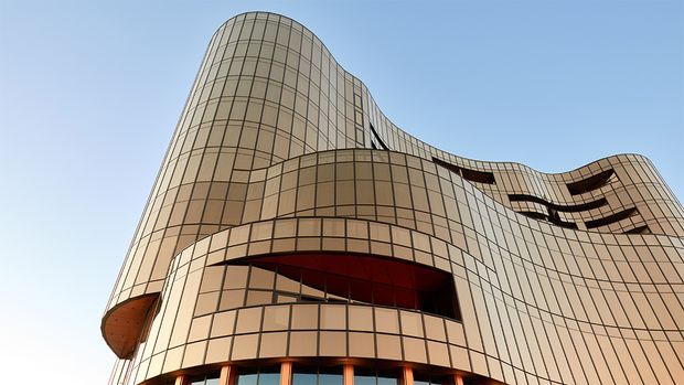 Low-angle exterior view of a large contemporary commercial building with a flowing, sculptural facade. The envelope is composed of curved bronze-beige glazed panels arranged in a gridded curtain-wall pattern, with recessed dark openings and a dramatic undulating roofline against a clear sky.