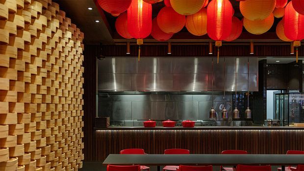 A warmly lit commercial restaurant interior, likely an Asian-inspired dining space, featuring a dramatic sculptural wall of stacked light timber blocks on the left, a stainless-steel open kitchen and extraction hood at the rear, dark vertical timber wall cladding, and a long counter with red stools. Numerous red and yellow paper lanterns hang across the ceiling, creating a vivid decorative canopy.