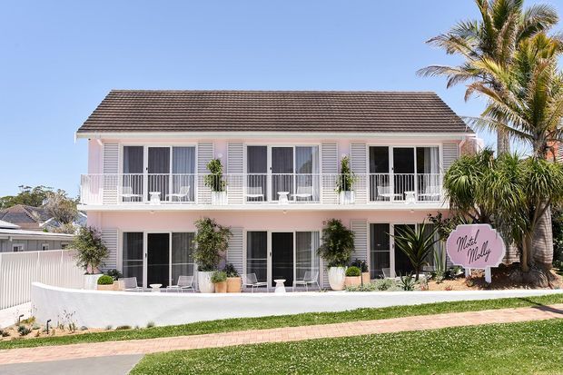 Front exterior view of a two-storey pale pink guesthouse or hotel-style building with a long tiled pitched roof, white shutters, extensive upper-level balconies, and ground-floor French doors. Tropical palms and landscaped planting frame the facade, with a paved pedestrian path and lawn in the foreground.