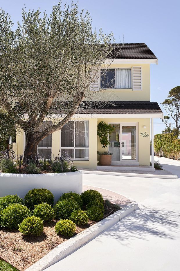 Bright exterior view of a two-storey suburban townhouse or villa with a pale cream rendered facade, dark tiled pitched roof, white-framed windows, louvred shutters, and a glazed entry door. The property is set behind manicured rounded shrubs, a mature olive tree, and a light concrete driveway and path.