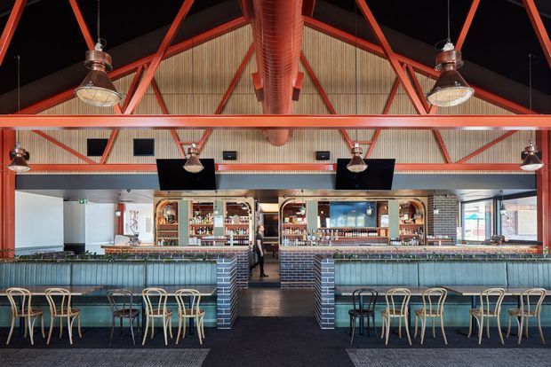 Interior view of a spacious contemporary commercial bar or hospitality venue. The room features a long tiled bar counter with teal upholstered frontage, timber bar stools and chairs, exposed red-painted steel roof trusses, corrugated metal ceiling panels, large cylindrical exposed ductwork, pendant lighting, dark flooring, and extensive back-bar shelving with bottles. The symmetrical composition and industrial structural detailing create a bold, lively hospitality setting.