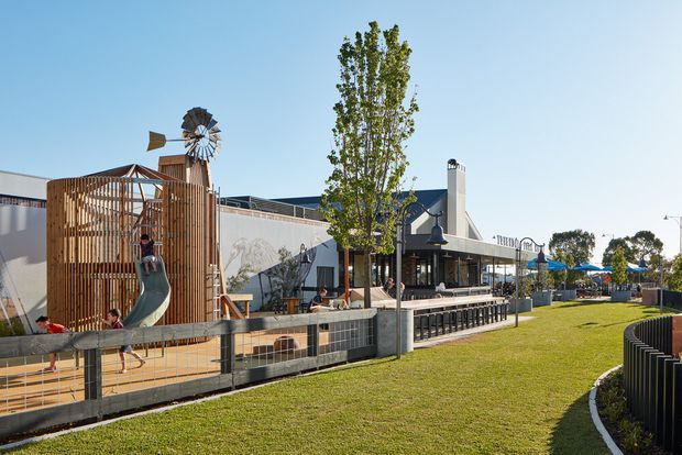 Exterior view of a contemporary commercial or community development with a fenced children's play area, sculptural timber screening, a small windmill feature, paved outdoor seating, and blue umbrellas. A landscaped grass verge and curved pedestrian edge lead toward the building frontage.