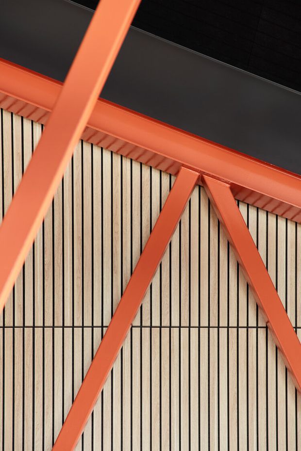 Close-up architectural detail of a bold orange-red painted steel structural frame with diagonal bracing, set against vertically ribbed light timber wall cladding and a dark ceiling or roof surface. The image emphasizes exposed structure, geometric composition, and contrasting industrial materials.