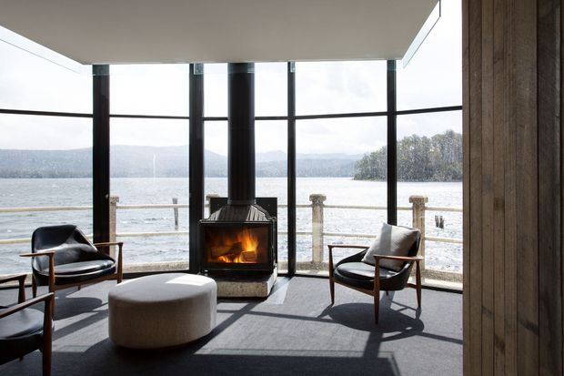 A refined modern living room with floor-to-ceiling glazing overlooking a broad body of water and distant hills. A freestanding wood-burning fireplace anchors the room, while mid-century-style lounge chairs and a rounded upholstered ottoman sit on a dark carpet. Vertical timber wall cladding frames the right side, and strong daylight creates a calm, scenic interior.