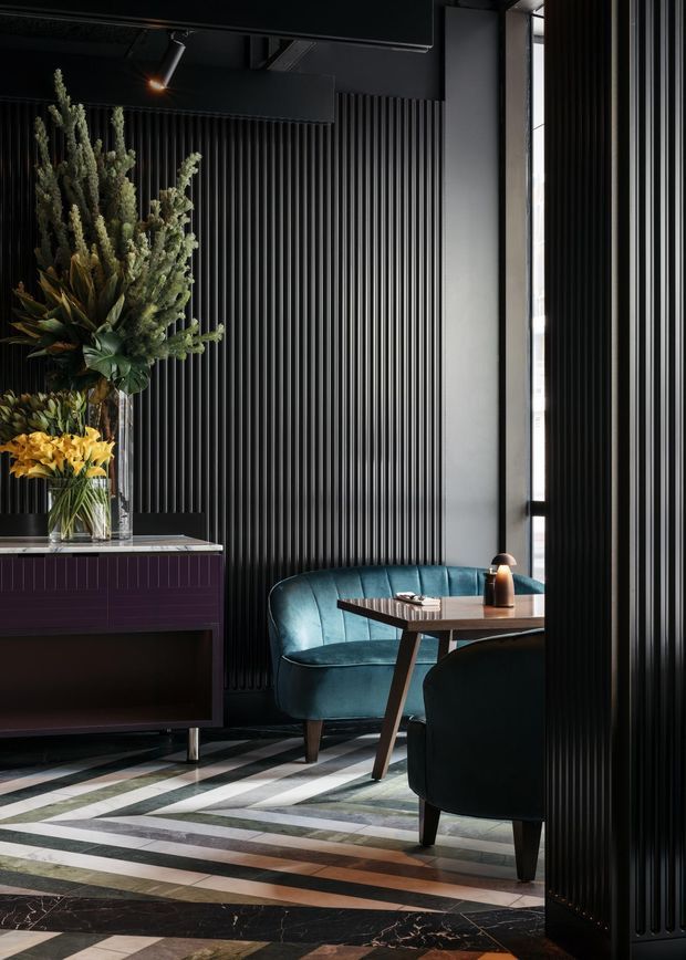 A moody, refined interior seating area with a teal curved sofa and matching dark armchair around a compact timber table. The room features dramatic black fluted wall panelling, a dark built-in sideboard with a stone top, tall narrow glazing, and a striking geometric floor composed of contrasting dark stone and timber-like bands. Sculptural floral and foliage arrangements add an organic accent, while a focused pendant or spotlight provides warm illumination.