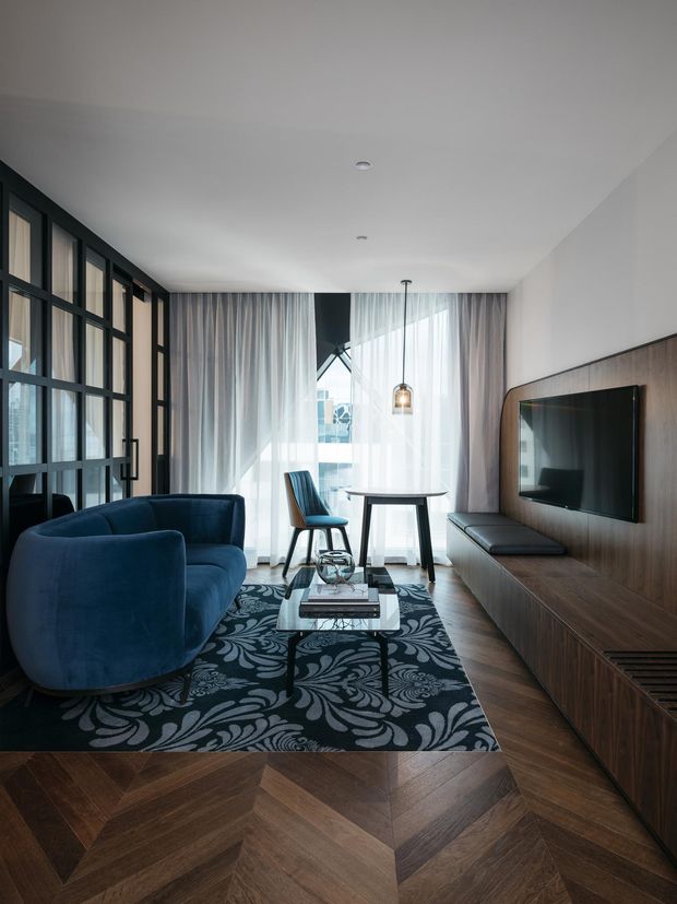 A refined contemporary living room with deep blue upholstered seating, a patterned dark rug, a glass coffee table, and a wall-mounted television integrated into a long dark timber media cabinet. Full-height sheer curtains frame bright glazing, while black-framed internal glazing separates the room from an adjacent space. The floor features a distinctive dark timber herringbone/parquet pattern, and a compact dining or work table sits by the window.