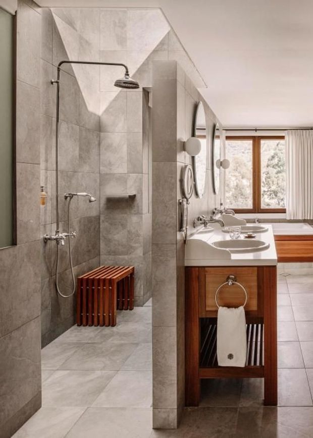 A refined bathroom with a walk-in shower clad in large grey stone-look tiles, exposed chrome shower fittings, a timber slatted bench, and a warm timber vanity with white vessel basins. Round mirrors and globe wall lights line the vanity, while a timber-framed window provides natural light. The space has a warm, understated contemporary character with strong natural materials.