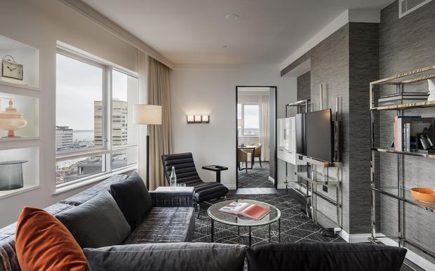 A stylish contemporary living room in an urban apartment, featuring a large dark sectional sofa, black leather lounge chair, glass coffee table, built-in display shelving, a wall-mounted television, and dark textured feature walls. Floor-to-ceiling windows provide city views, while neutral furnishings and warm lighting create a refined, comfortable atmosphere.